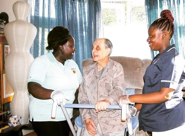 Two Muzaana carers supporting an older woman as she walks with a frame at home.